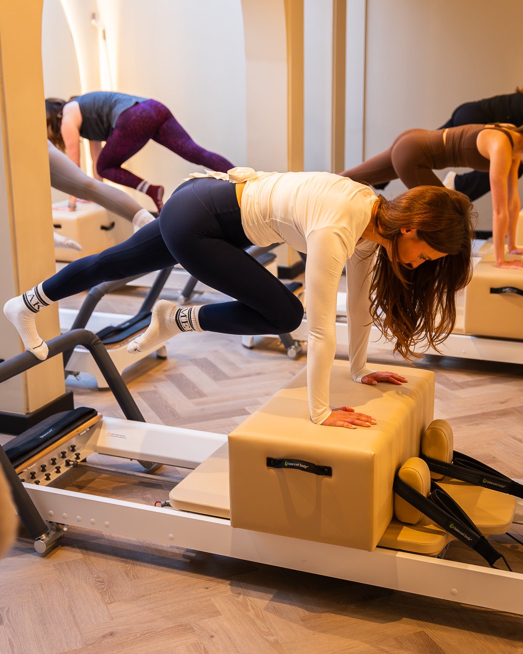 Women exercising on Pilates reformer machine and balance cushion in a fitness studio.