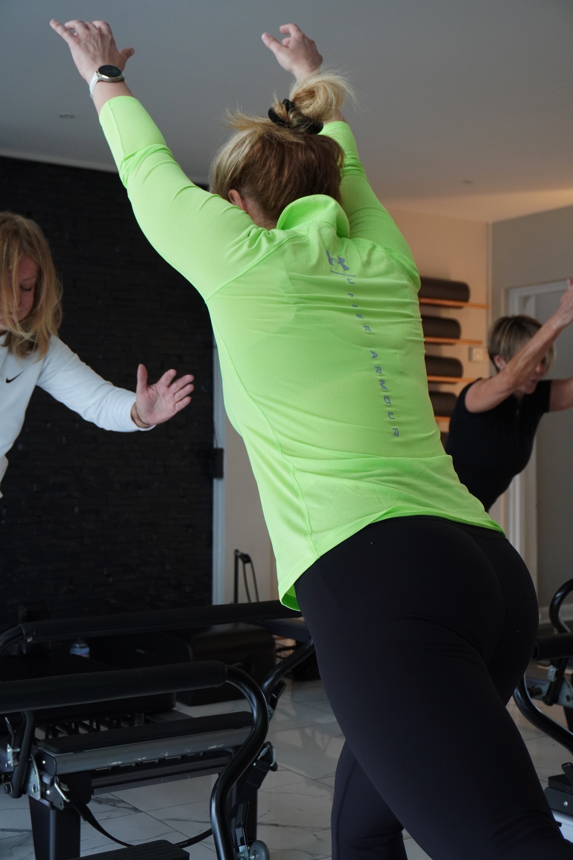 Three women exercising in a fitness class, stretching with their arms raised overhead.