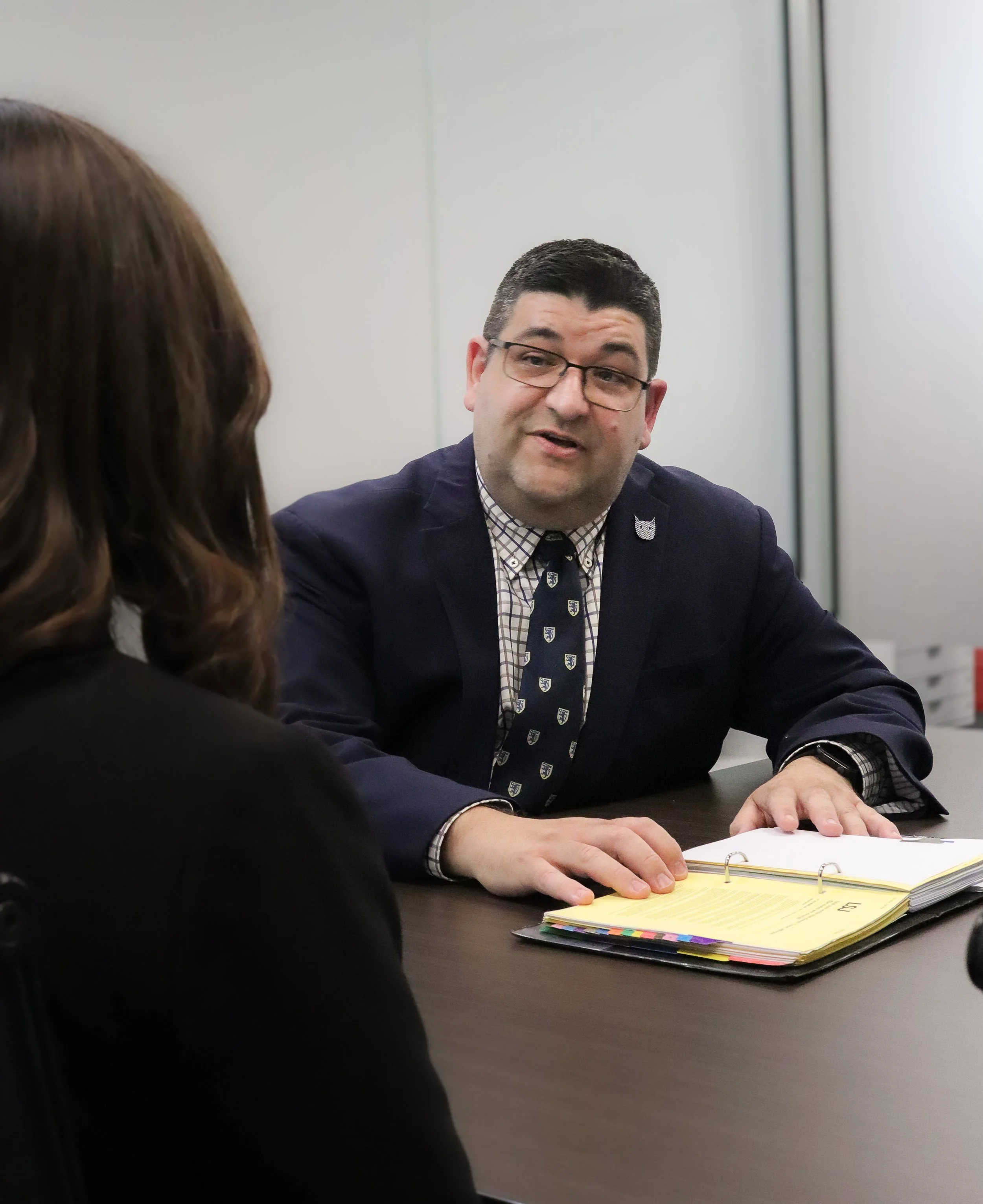 A man in a suit and glasses sitting at a desk, talking to a woman with brown hair, with a folder of papers in front of him.