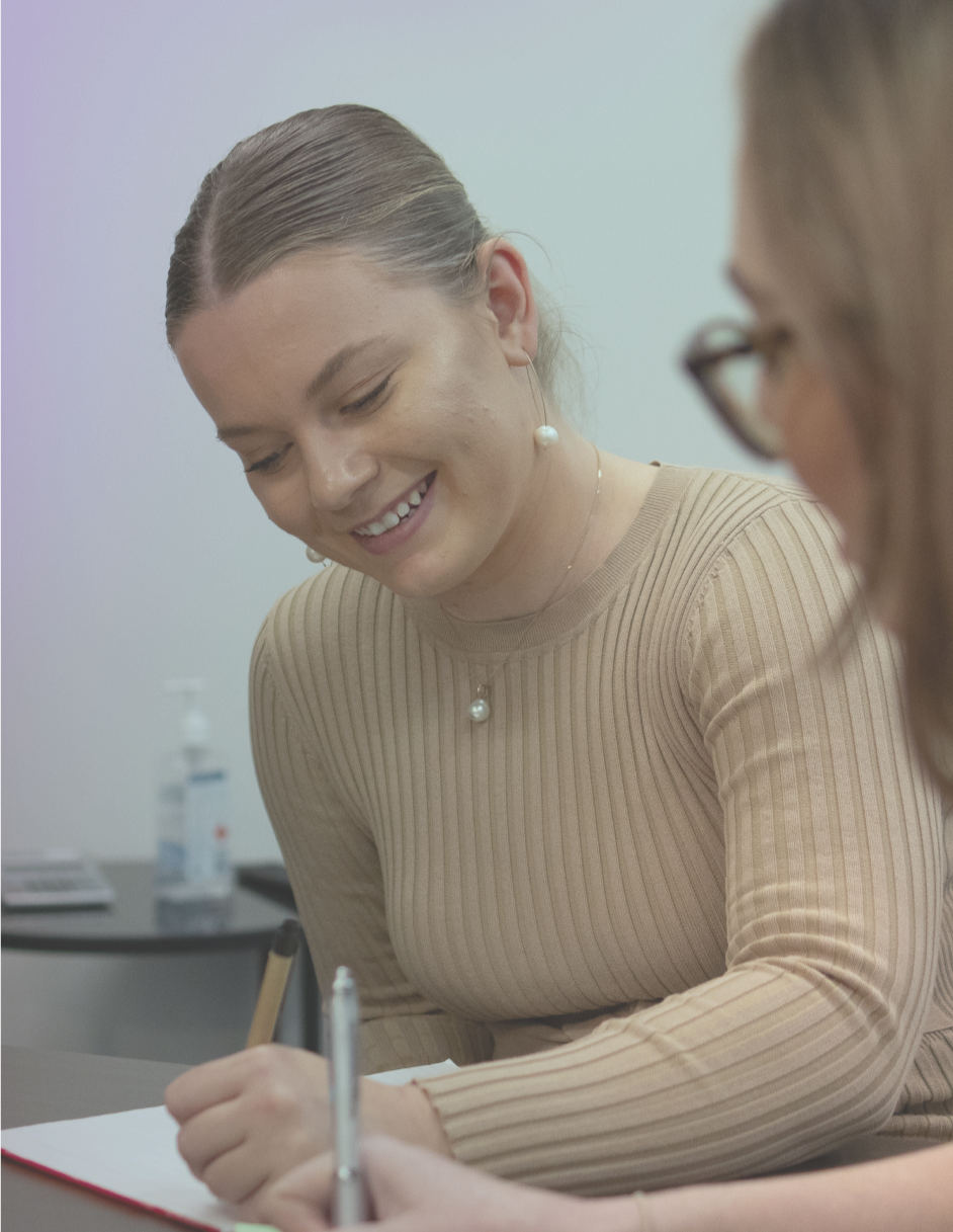 A woman with brown hair tied back, wearing a beige ribbed long-sleeve top, pearl earrings, and a matching pearl necklace, smiling and writing on a notebook while seated at a table during a meeting.