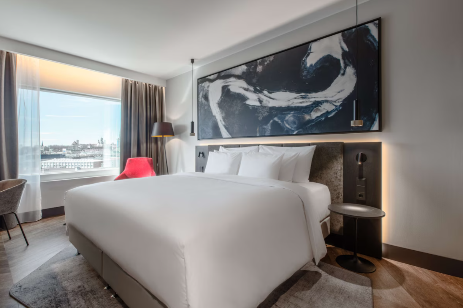 Modern hotel room with a large bed, abstract artwork above, window with city view, and stylish lighting.