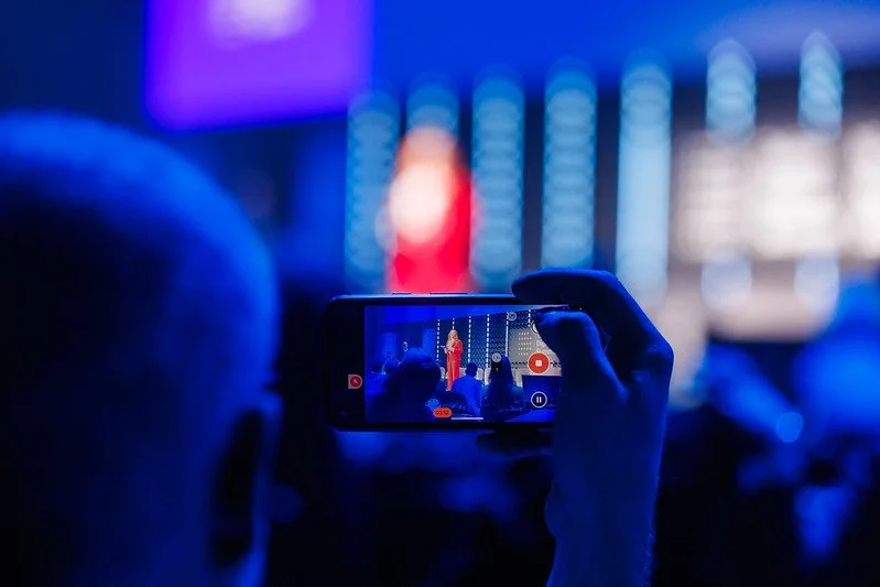 Person recording a speaker on stage with a smartphone at an event.
