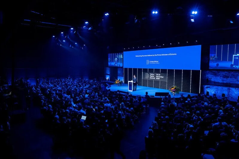 A large conference or event hall filled with seated audience facing a stage with a speaker at a podium. Large screens behind the speaker display a blue presentation slide with text welcoming the Prime Minister of Estonia. The room is dimly lit with blue lighting and floral arrangements on the stage.