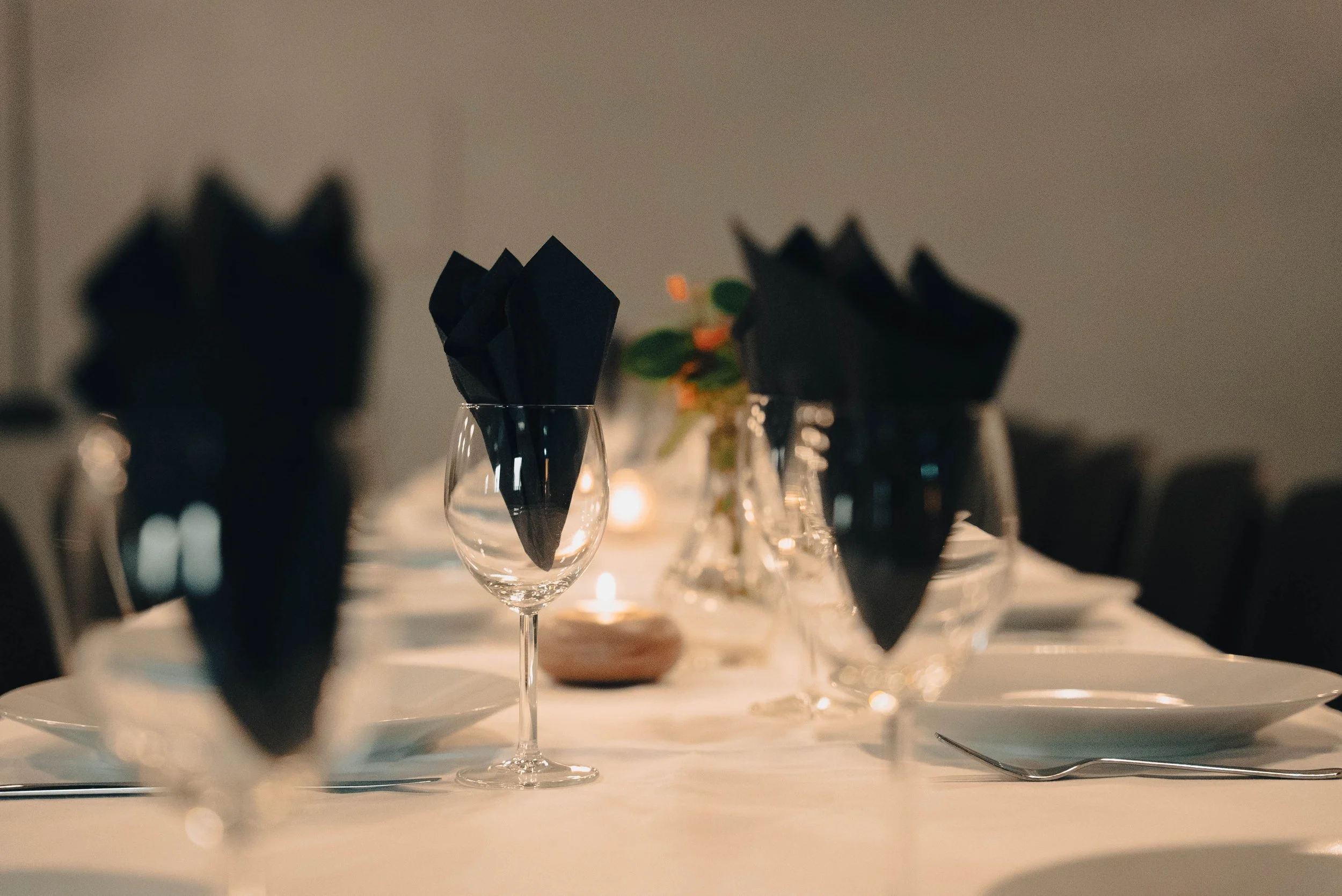 Klar til middagsbord med vin glass, sorte papirservietter, tallerkener og blomsterdekorasjon i bakgrunnen, romantisk stemning.