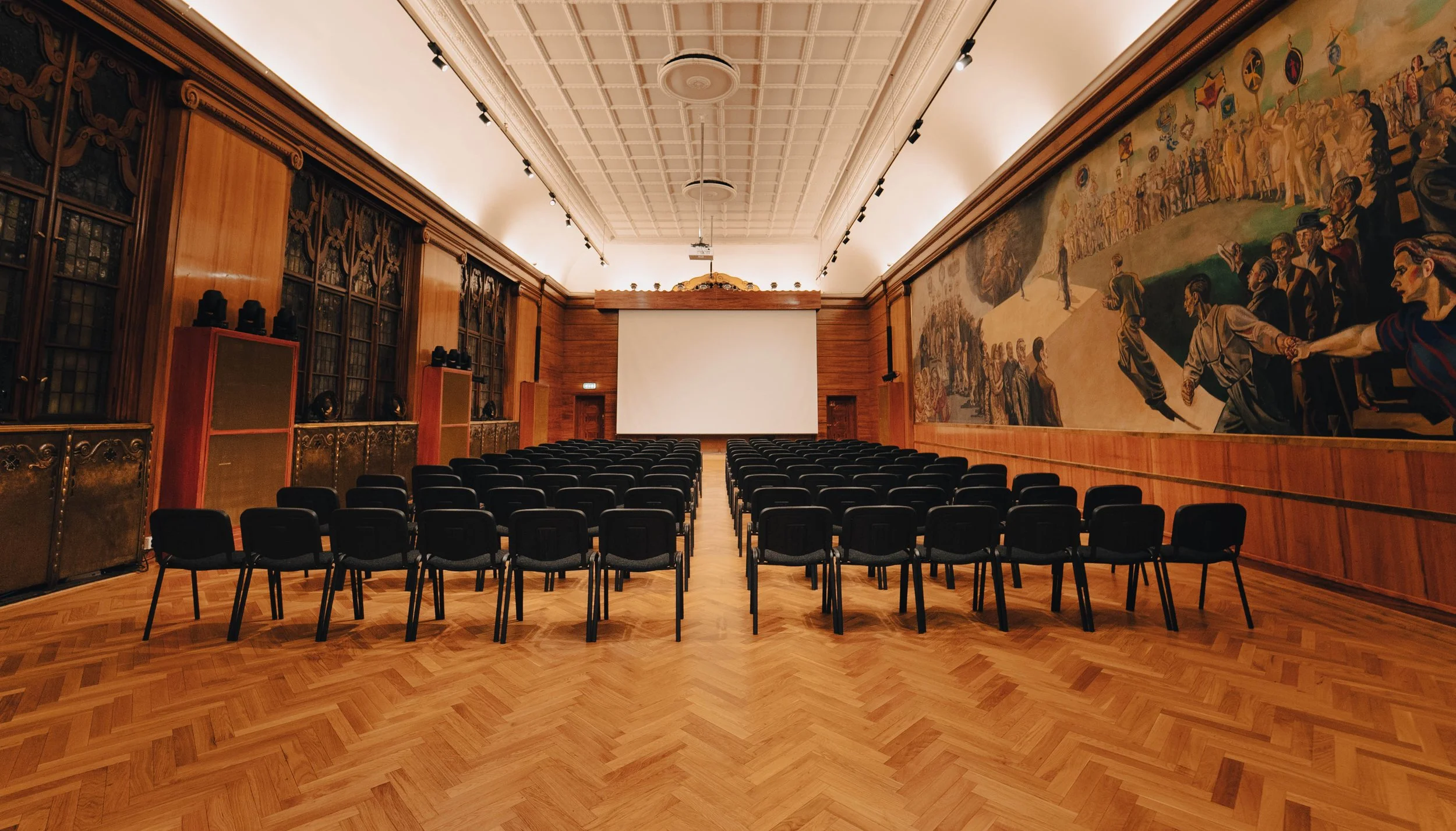 Stort tomt auditorium med rader av svarte stoler, en stor projeksjonsskjerm og en lang veggdekorasjon med historiske bilder på treverk.