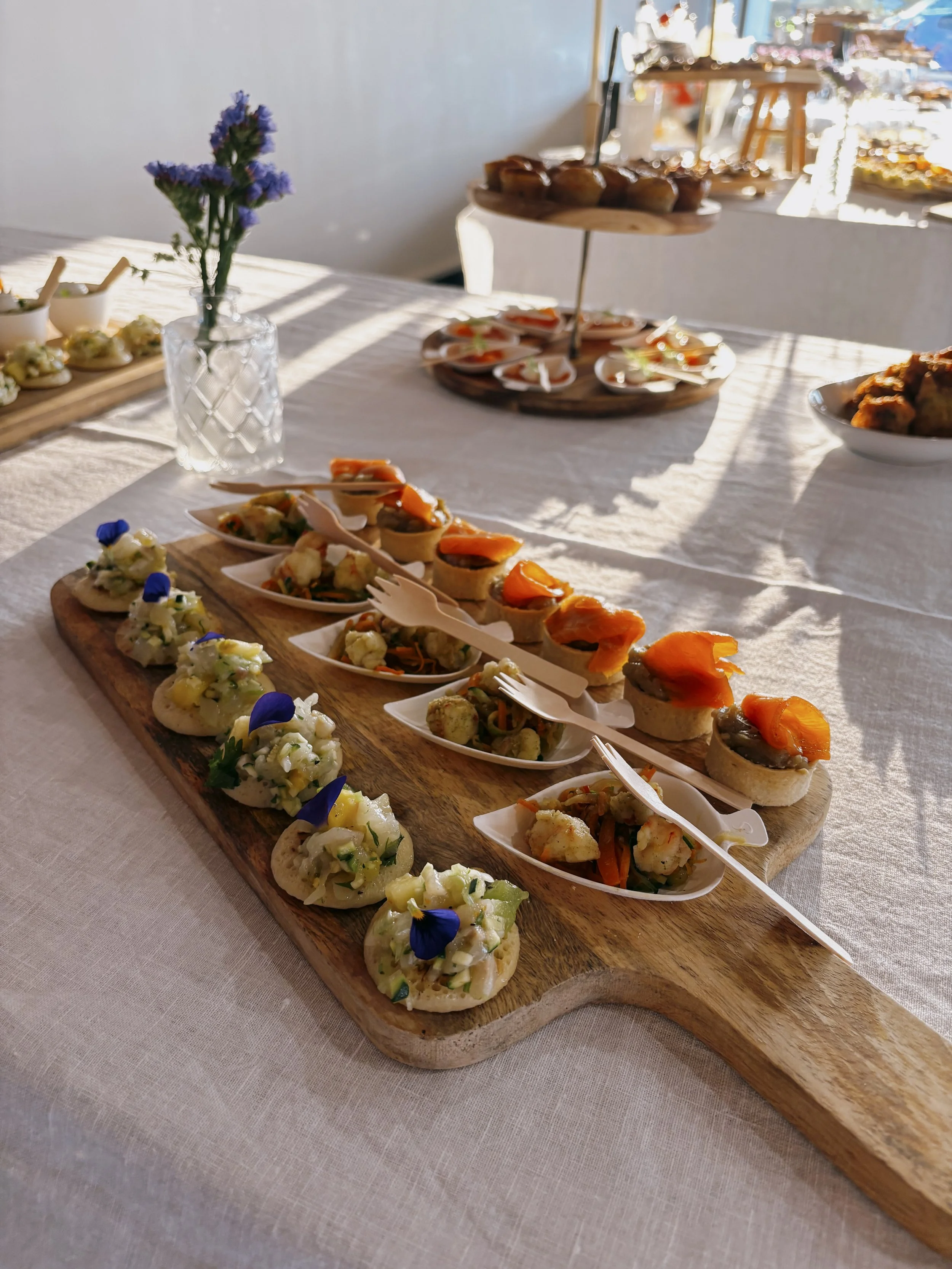 Assortiment de petites bouchées et amuse-gueules sur un plateau en bois, décoré de fleurs violettes dans un vase en verre, à une table avec nappe blanche, en ambiance lumineuse et chaleureuse.
