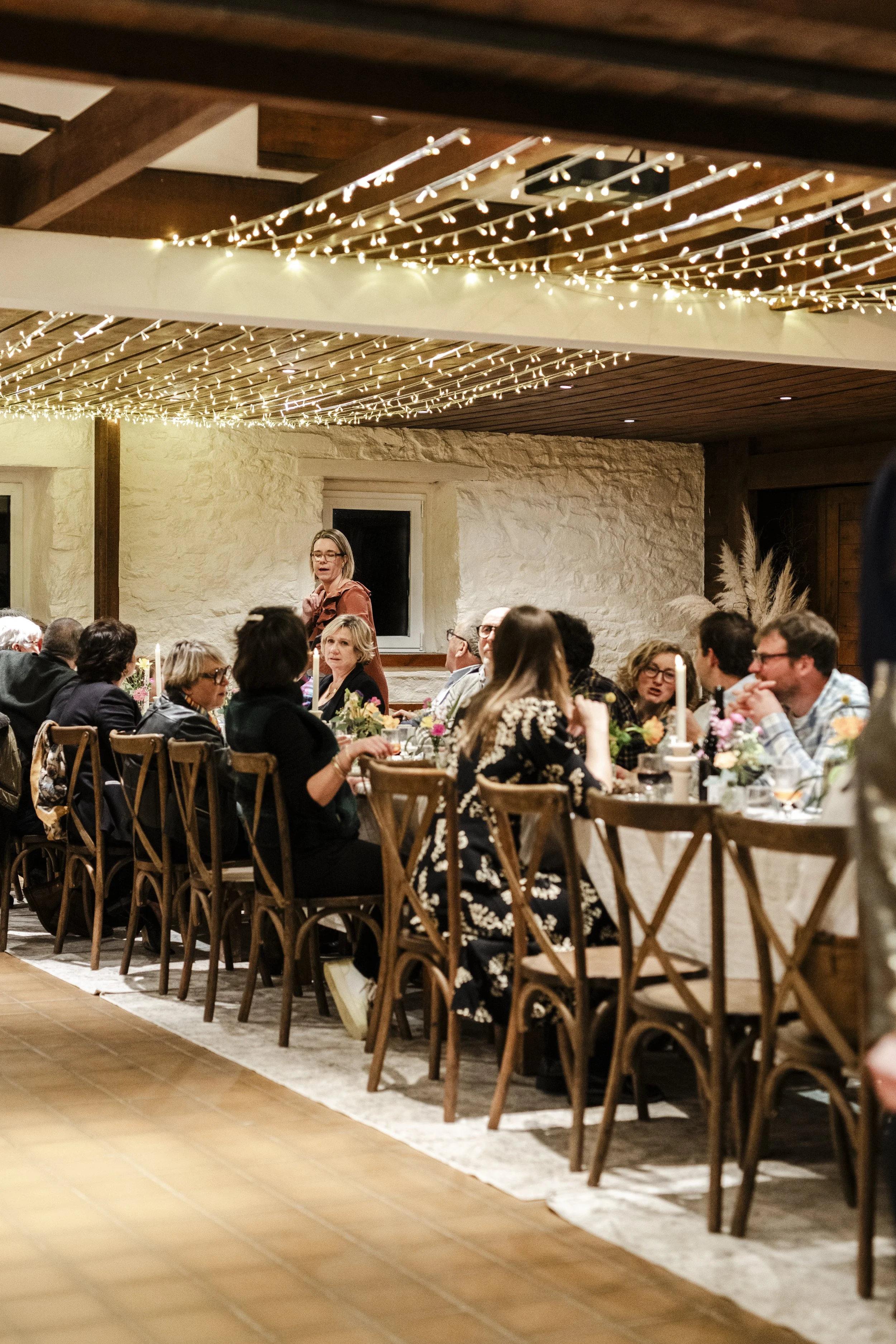 Groupe de personnes assises à une longue table lors d'un repas ou d'une réception, décorée avec des fleurs et des bougies, sous un plafond orné de guirlandes lumineuses