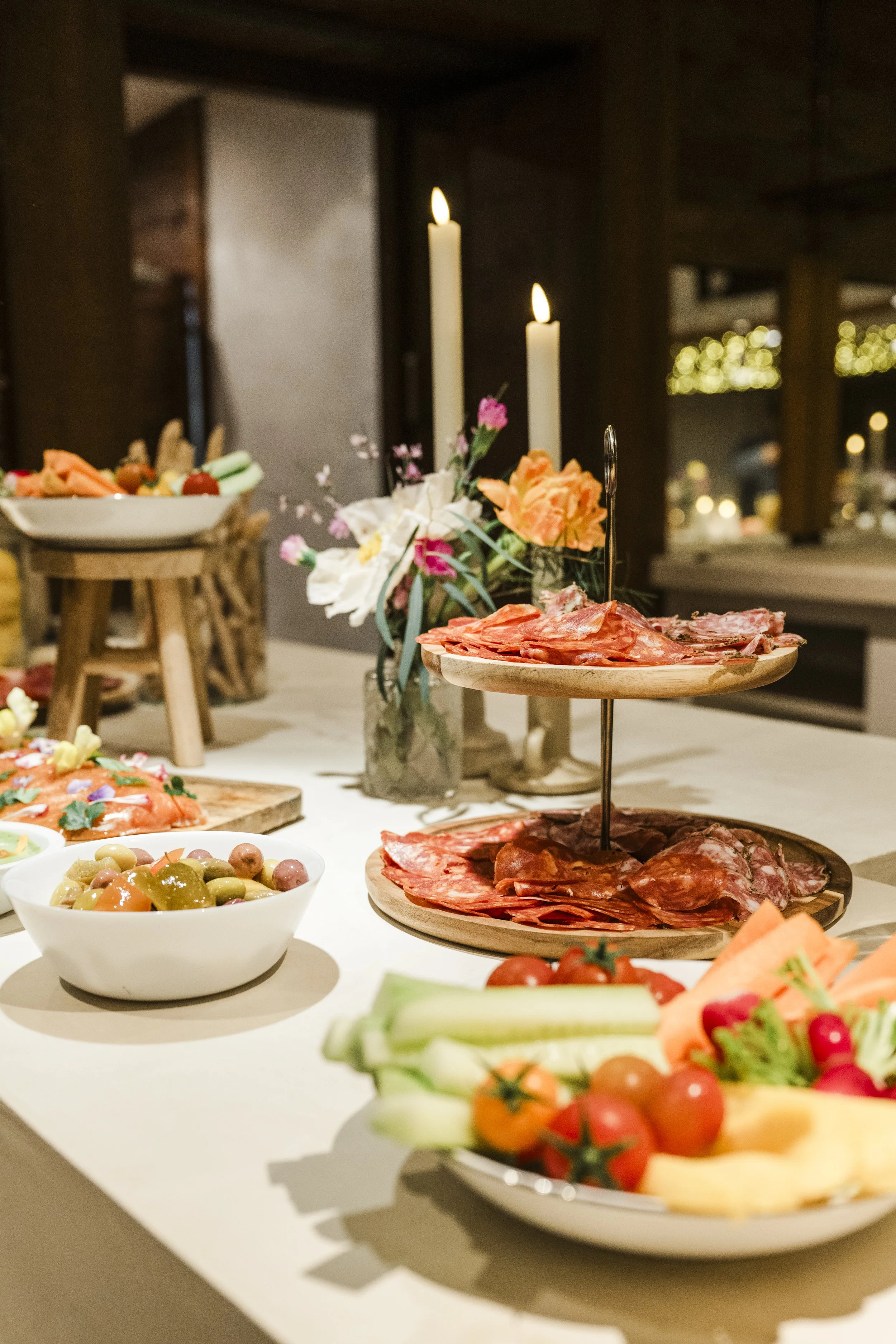 Plateau de charcuterie avec différentes tranches de viande, bol de tomates et légumes, fleurs, bougies allumées sur une table lors d'un repas.