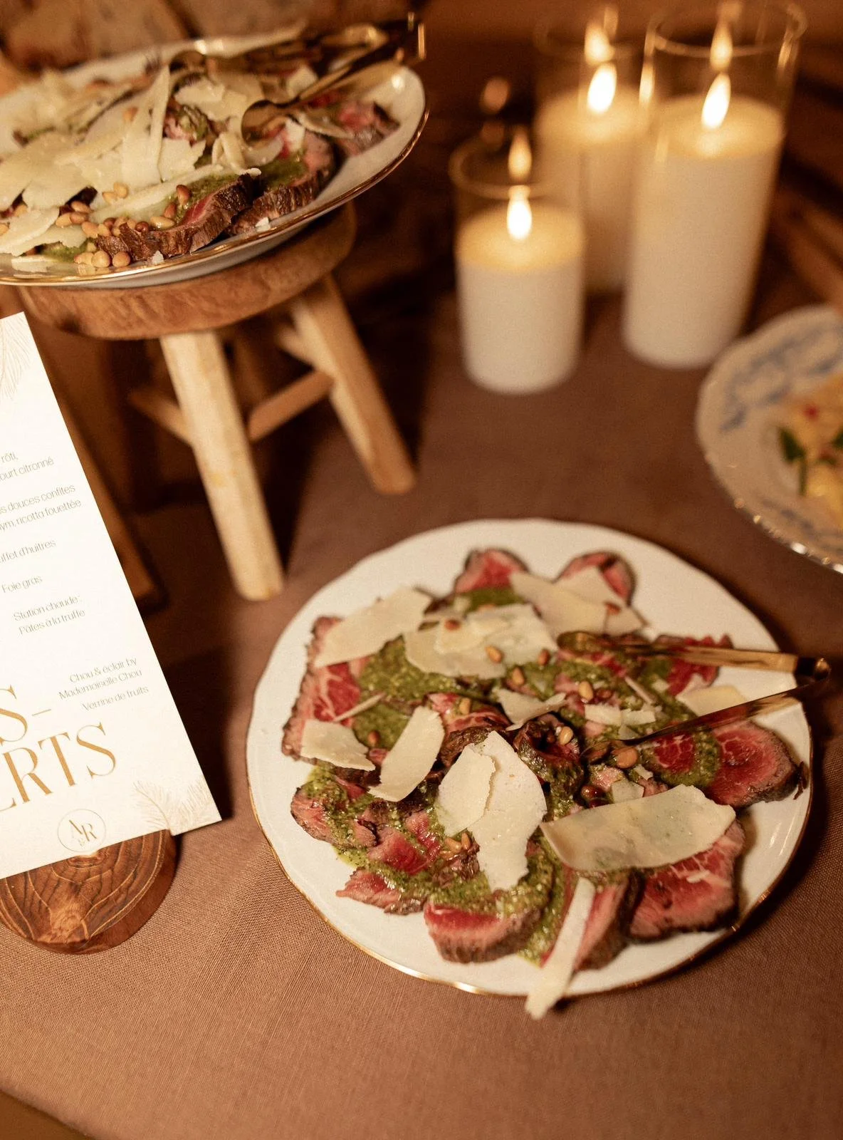 Une assiette avec du carpaccio de viande, des copeaux de fromage, du pesto, des graines ou noix. En arrière-plan, des bougies allumées et une autre assiette, créant une ambiance chaleureuse.