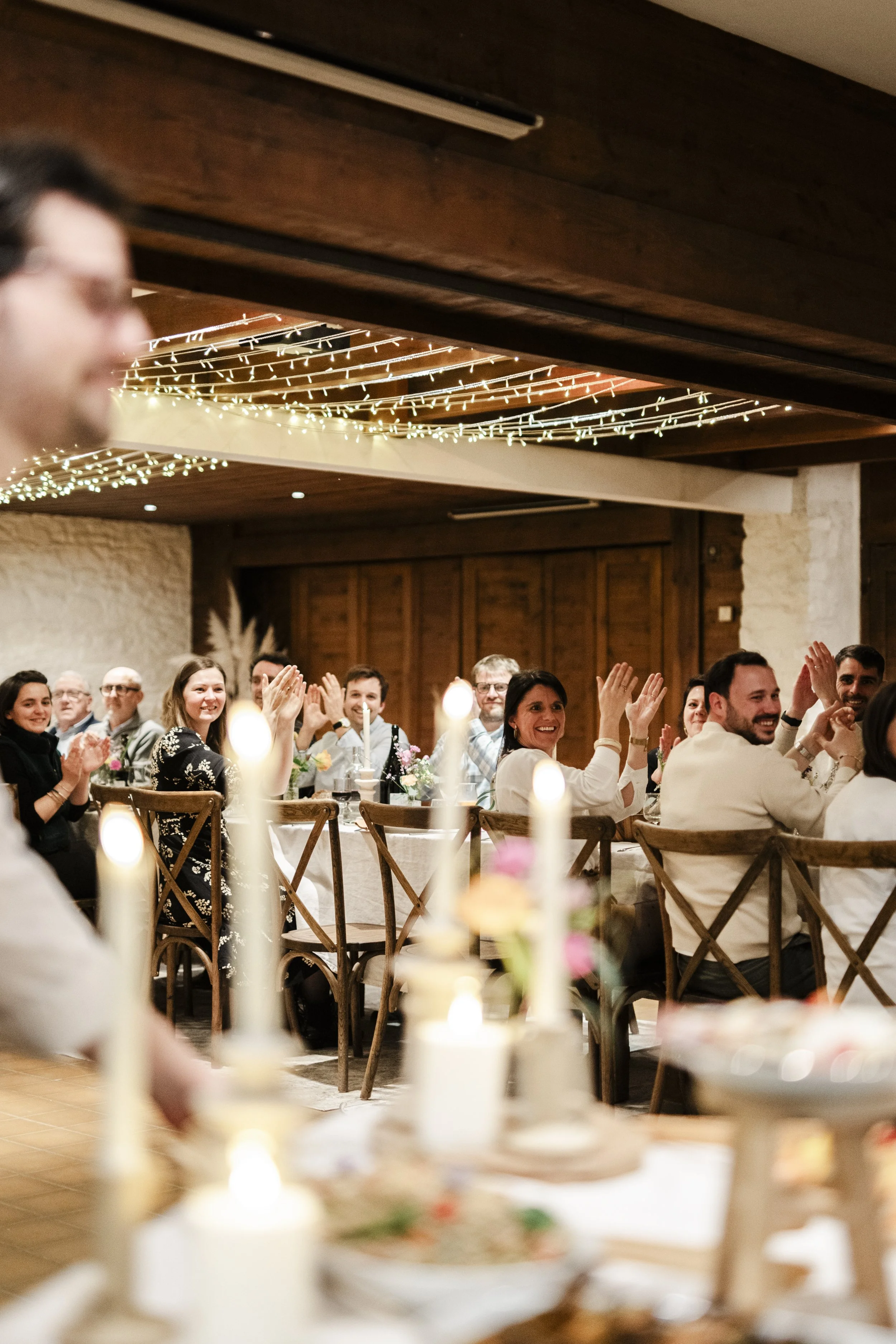 Groupe de personnes assises à une table lors d'une célébration, probablement un mariage, dans une salle chaleureuse avec des guirlandes lumineuses au plafond.
