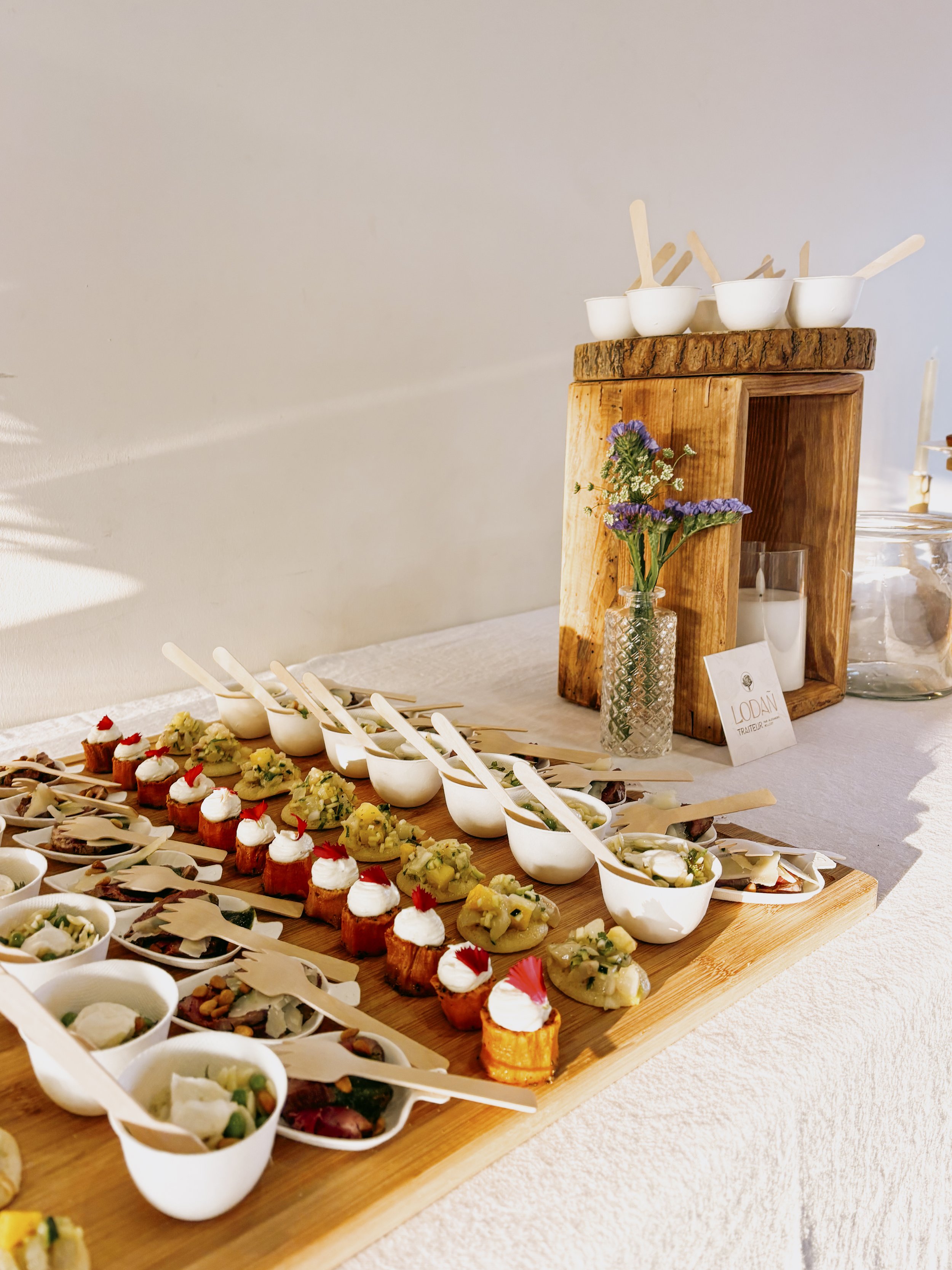 Plateau de dégustation avec des amuse-bouches et des entrées en portion individuelle, décoré avec une petite vase de fleurs et une caisse en bois en arrière-plan sur une table blanche.