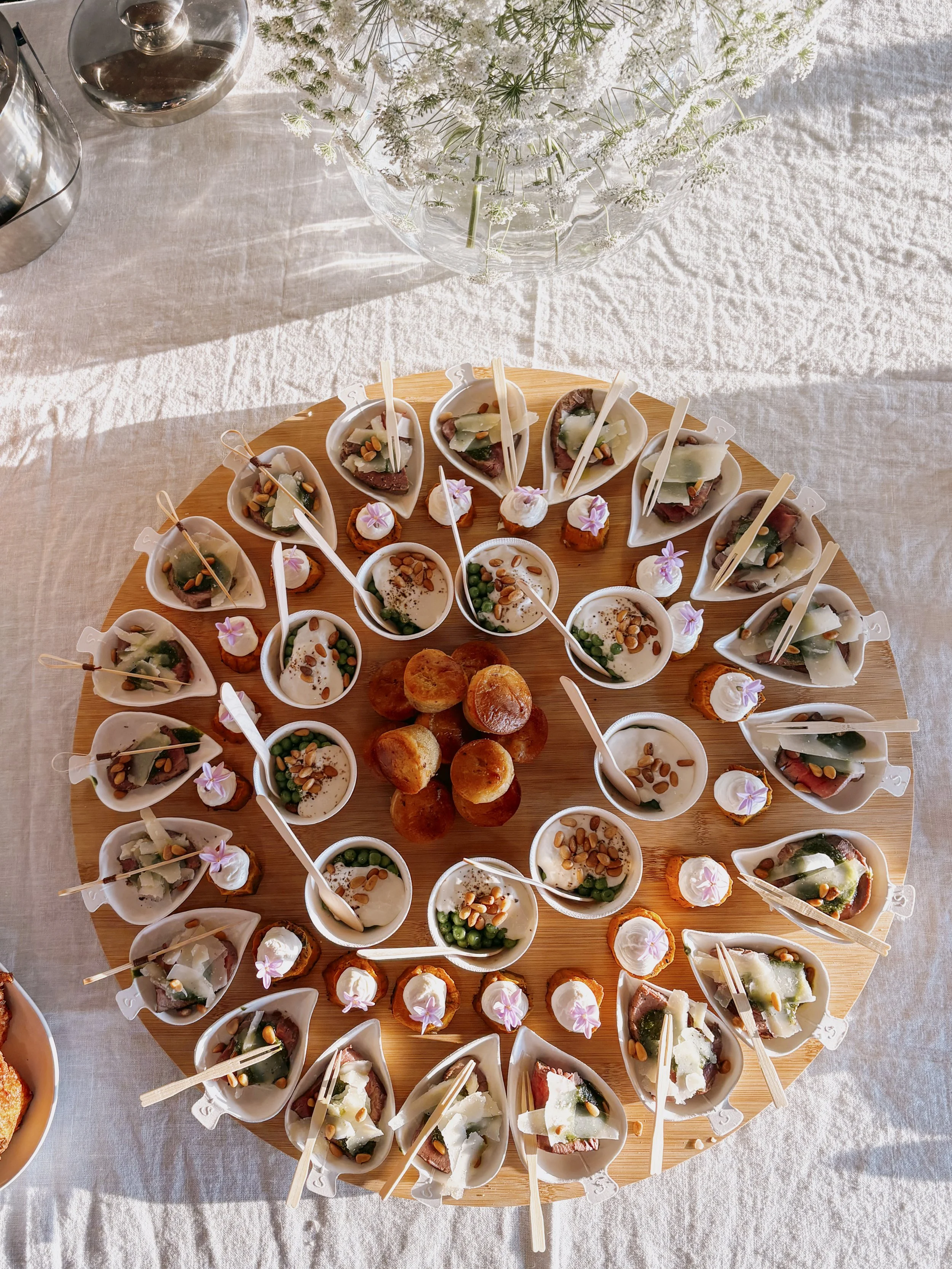 Plateau de miniature hors-d'œuvre avec diverses verrines et bouchées, décoré avec des fleurs, sur une nappe beige.