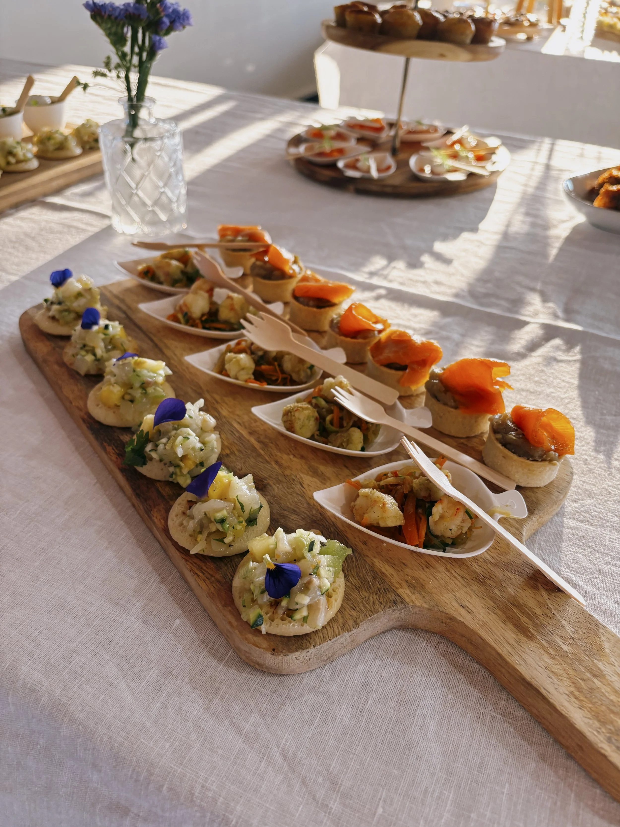 Plateau de amuse-gueules avec des canapés garnis de saumon, de légumes et de fleurs comestibles, sur une planche en bois, avec une assiette de mini sandwiches en arrière-plan.