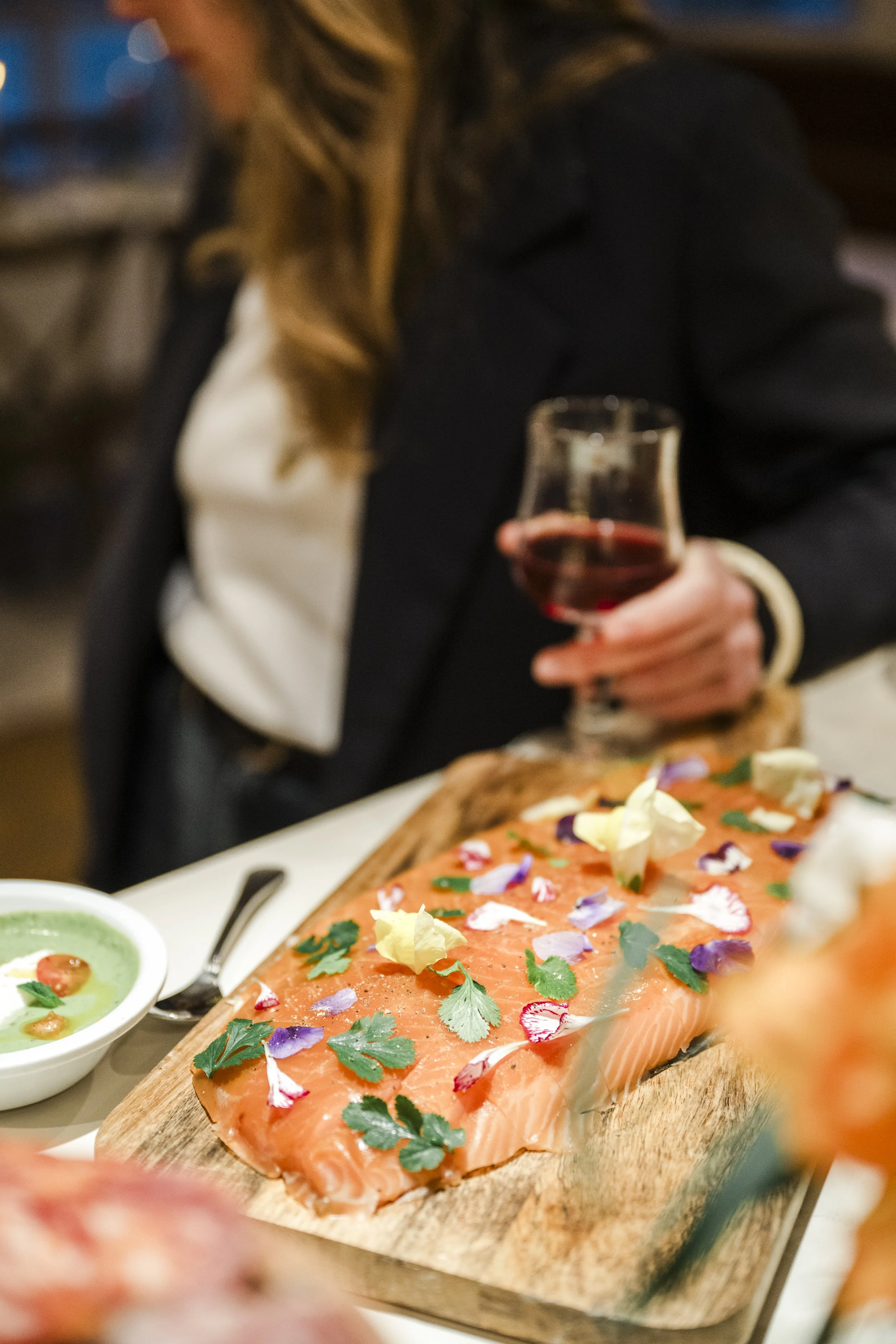 Fille tenant un verre de vin rouge à côté d'un plat de saumon décoré de fleurs et d'herbes, avec une soupe verte en arrière-plan.
