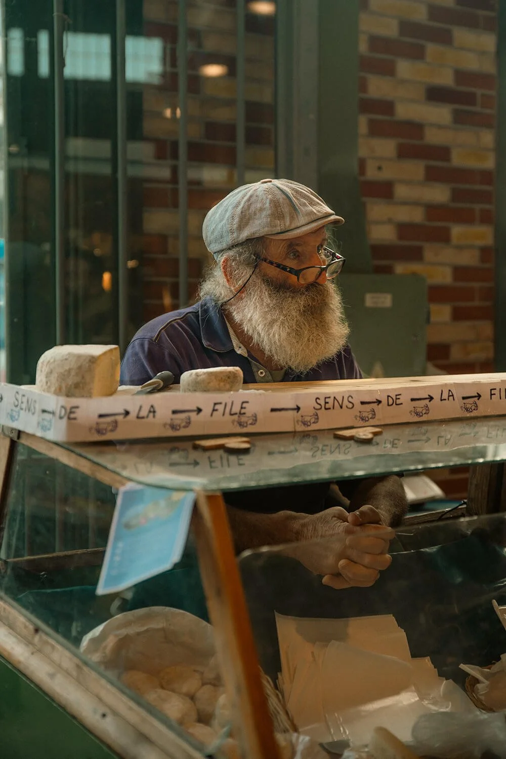 An elderly man with a gray beard, glasses, and a beige cap, sitting behind a cheese counter in a store, with a brick wall and glass window in the background. Taken in Rennes, France.