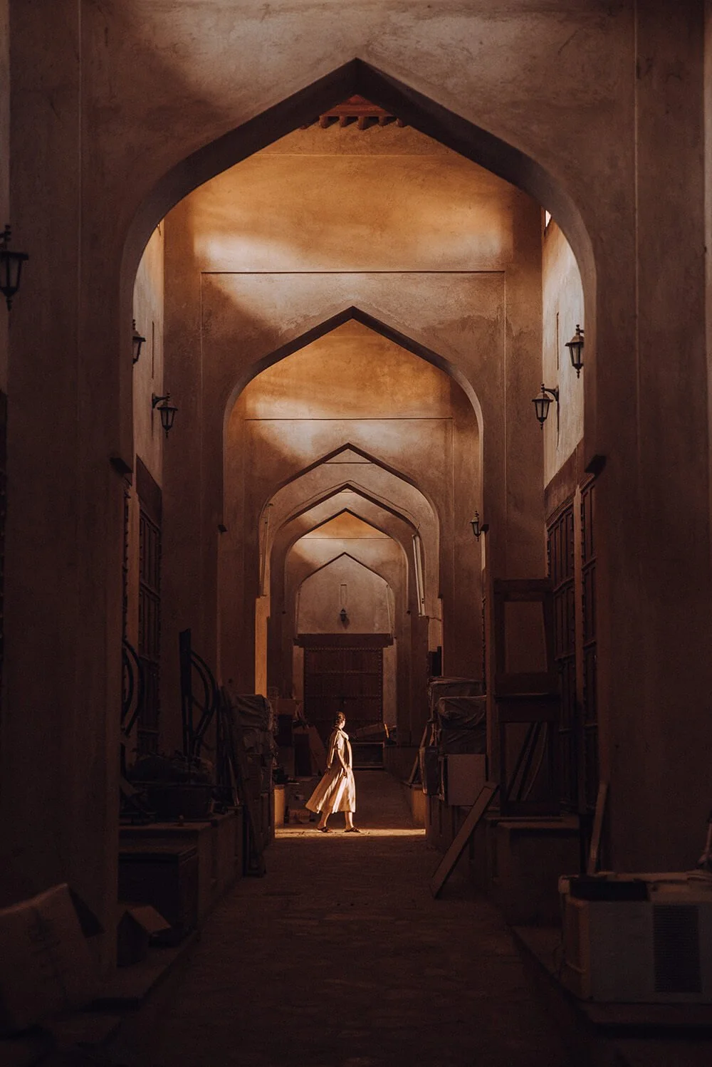 A woman walking through a historically styled, arched corridor with warm lighting and vintage furniture on either side. Taken in the Nizwa Souk in Oman.