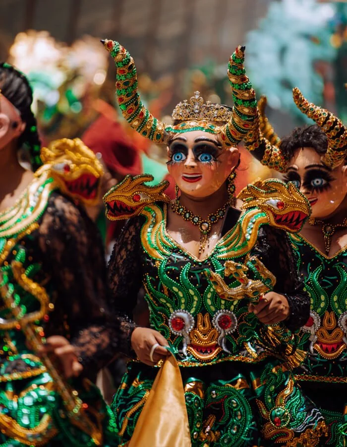 People in colorful, elaborate costumes with masks, headpieces, and face paint, participating in a cultural parade or celebration. Taken during the Carnival of Oruro in Bolivia.