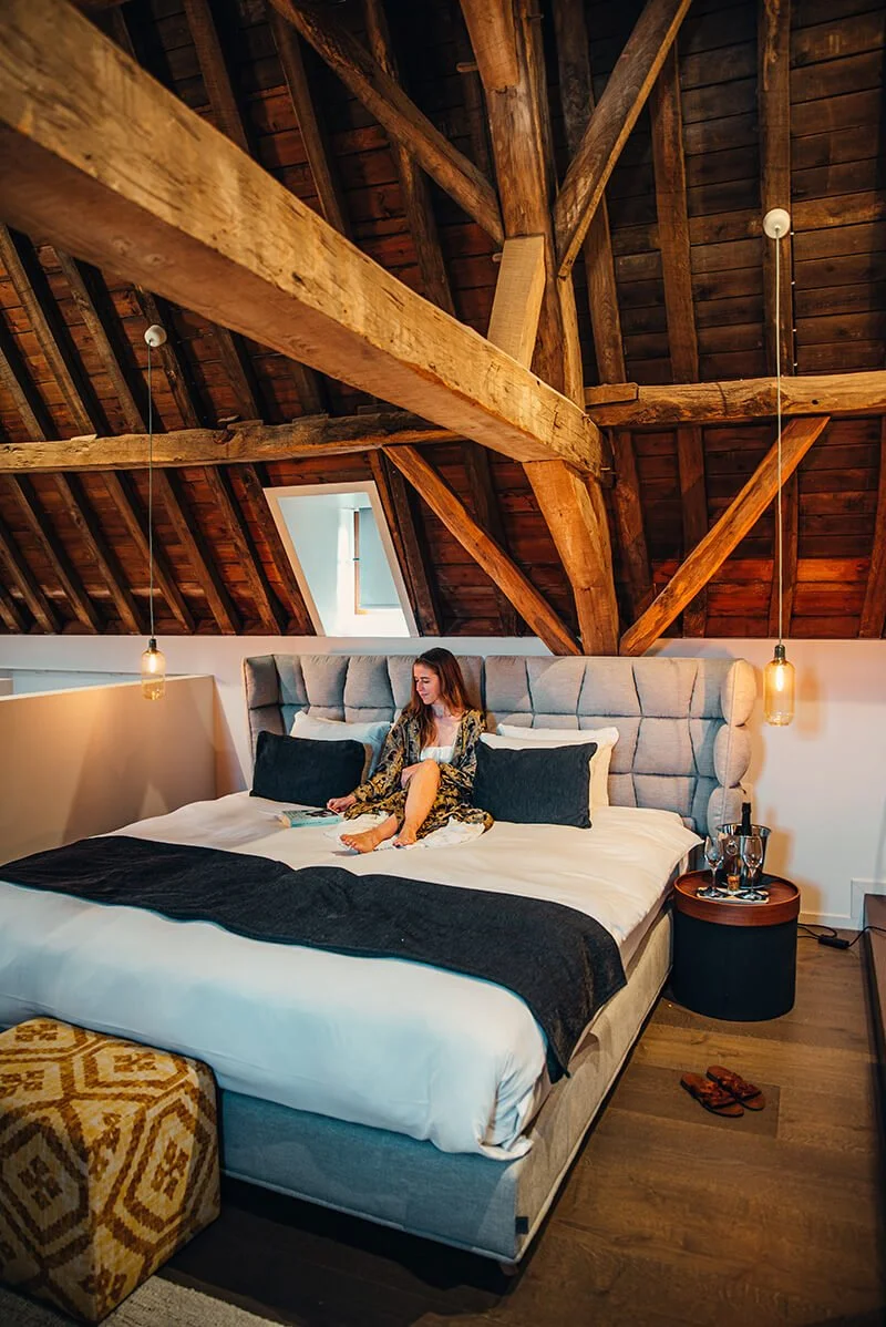 A woman sitting on a bed in a cozy attic bedroom with wooden beams and a small window. She is reading a book and wearing a patterned robe. The bed has a tall padded headboard, black throw pillows, and is covered with white linens and a black blanket.