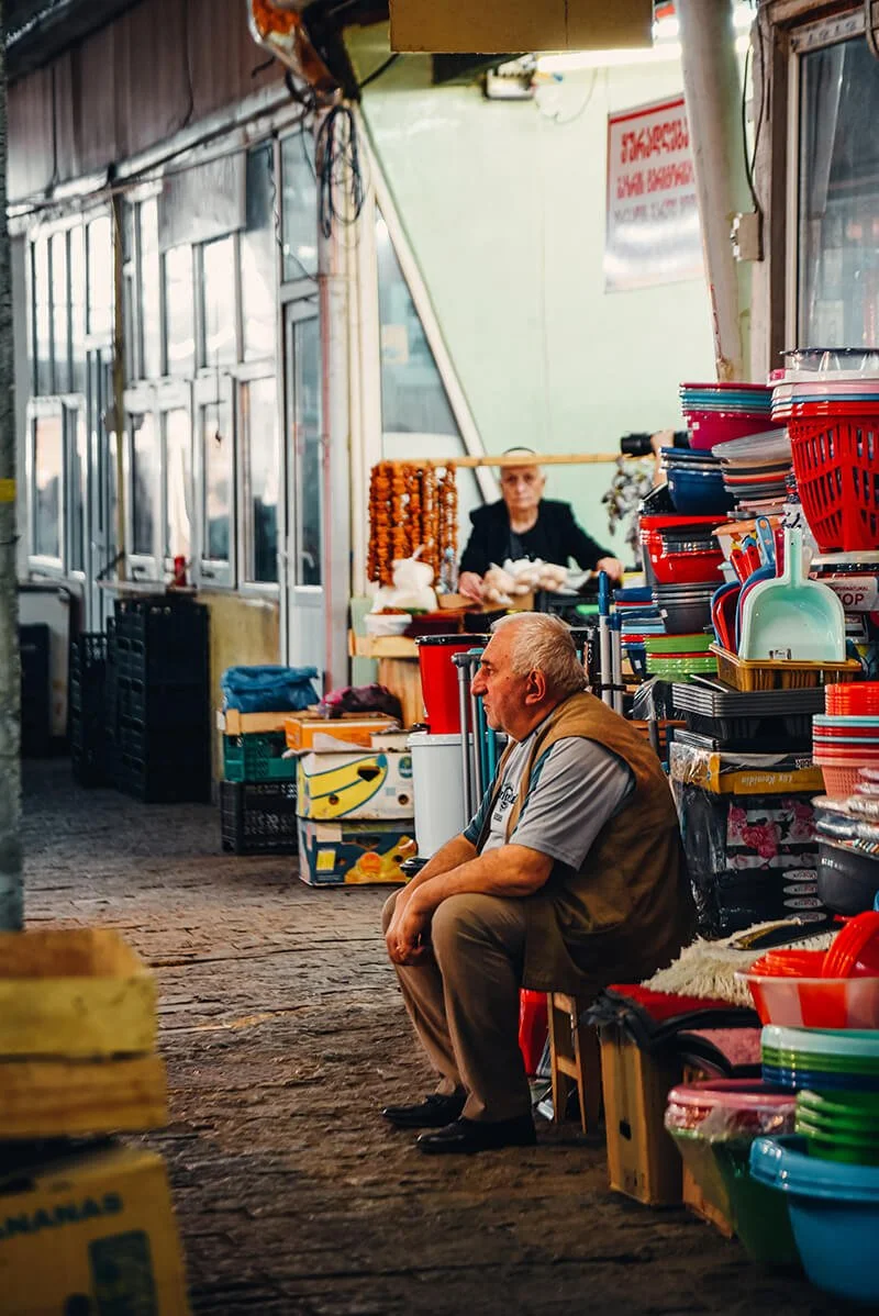 A man sitting on a small stool in a marketplace, surrounded by colorful plastic bowls and kitchenware stacked on shelves. In the background, a woman behind a stand with hanging garlic or onions. The marketplace has an earthy, rustic flooring with nat