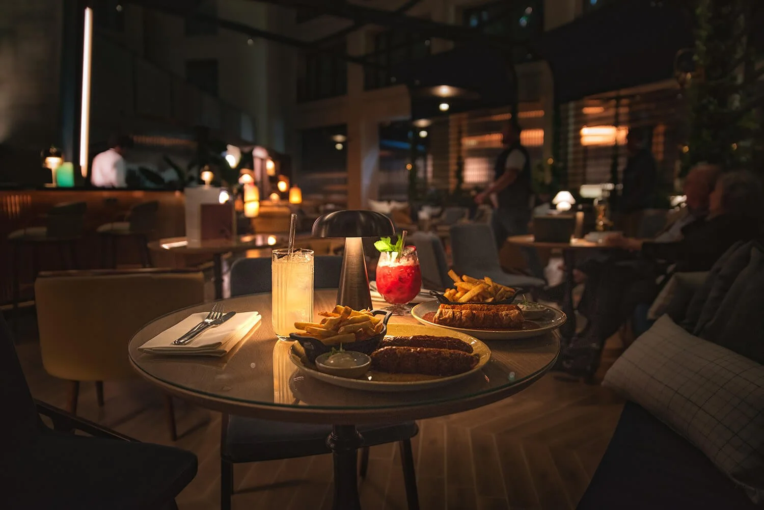 Restaurant table set with two plates of food, two drinks, and a candle in a cozy, dimly lit atmosphere with other diners in the background. Example of hotel UGC.