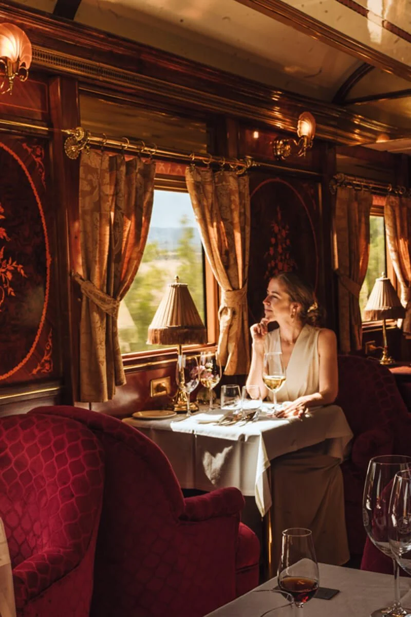 A woman in an elegant sleeveless dress sitting at a dining table in a luxurious train restaurant, with wine glasses and a table setting, looking out the window on a sunny day.