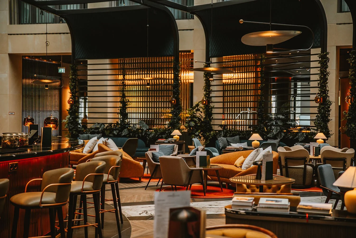 Interior of a stylish lounge or hotel lobby with modern furniture, lamps, and decorative greenery, illuminated by warm lighting.