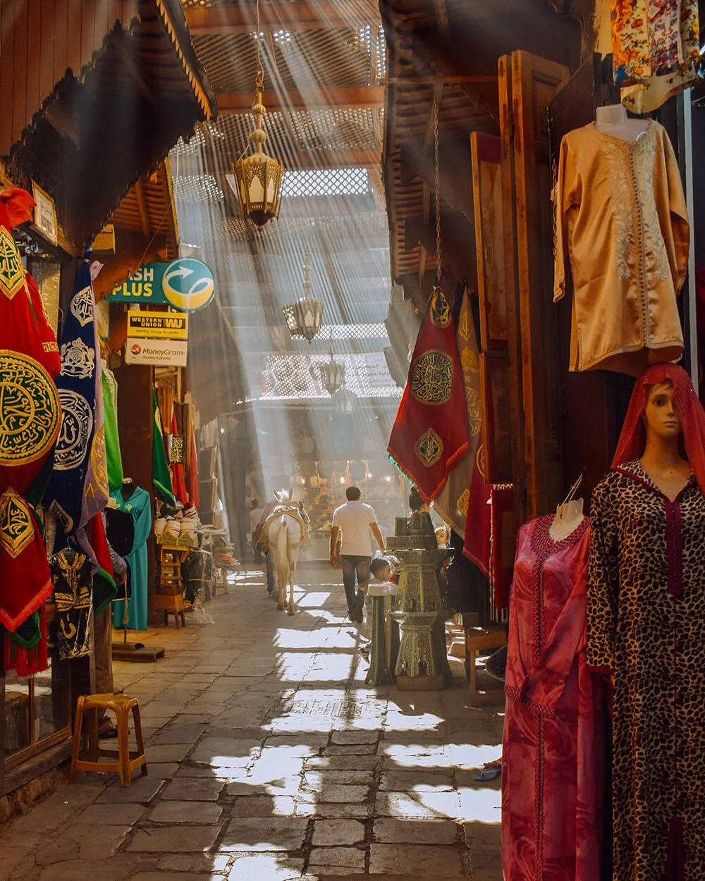 People walking through a vibrant market alley with colorful textiles and clothing on display on both sides, sunlight streaming through the roof. Taken in the Medina of Fez, Morocco.