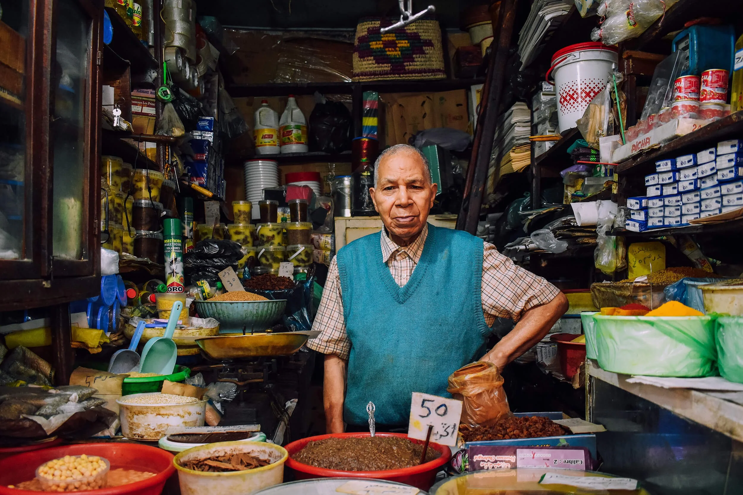 An elderly man in a blue vest stands behind a market stall filled with dried spices, herbs, and packaged goods, with shelves of jars, boxes, and bags in the background. Taken in the Medina of Fez, Morocco.