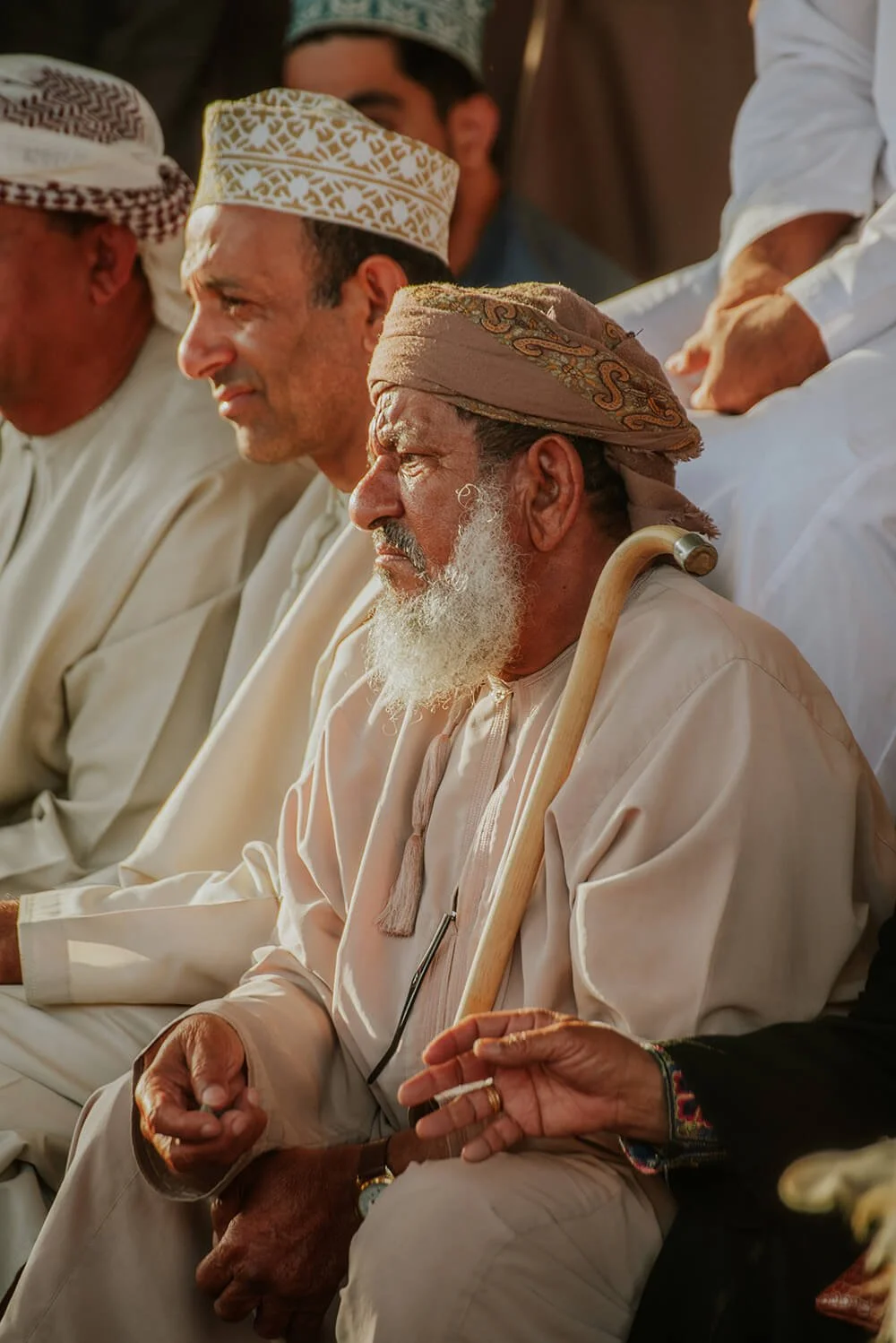 A group of Muslim men seated, dressed in traditional white clothing and head coverings, with one elderly man holding a cane and wearing a beige turban, during a gathering or event. Goat Market in Nizwa, Oman.