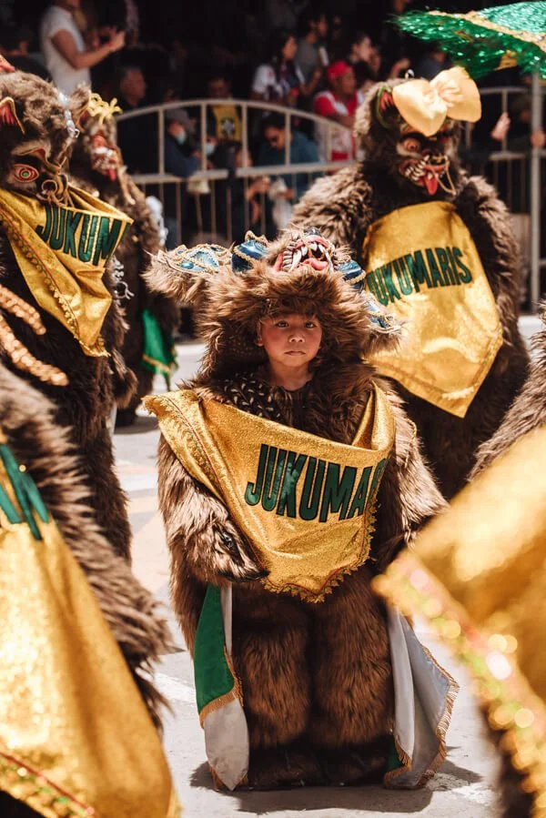Child dressed in traditional brown fur costume with a yellow sash that reads "JUKUMA" participating in a cultural parade, surrounded by other participants in similar costumes with masks and hats. Taken during Oruro Carnival in Bolivia.