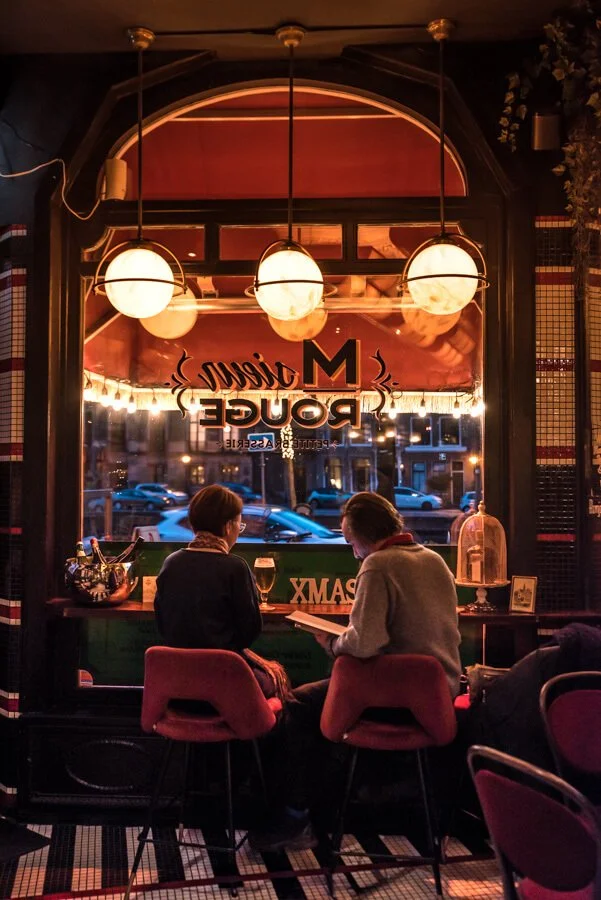 Two women sitting at a bar counter by a large window, with outdoor street view and parked cars; warm indoor lighting and Christmas decorations inside a cozy bar or restaurant. Taken in Haarlem, Netherlands.
