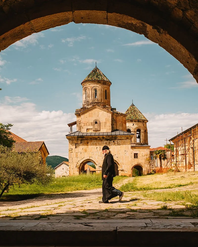 A person dressed in black walking past an old stone church with two domed towers, as seen through an arched stone window. Example of street photography in Georgia.