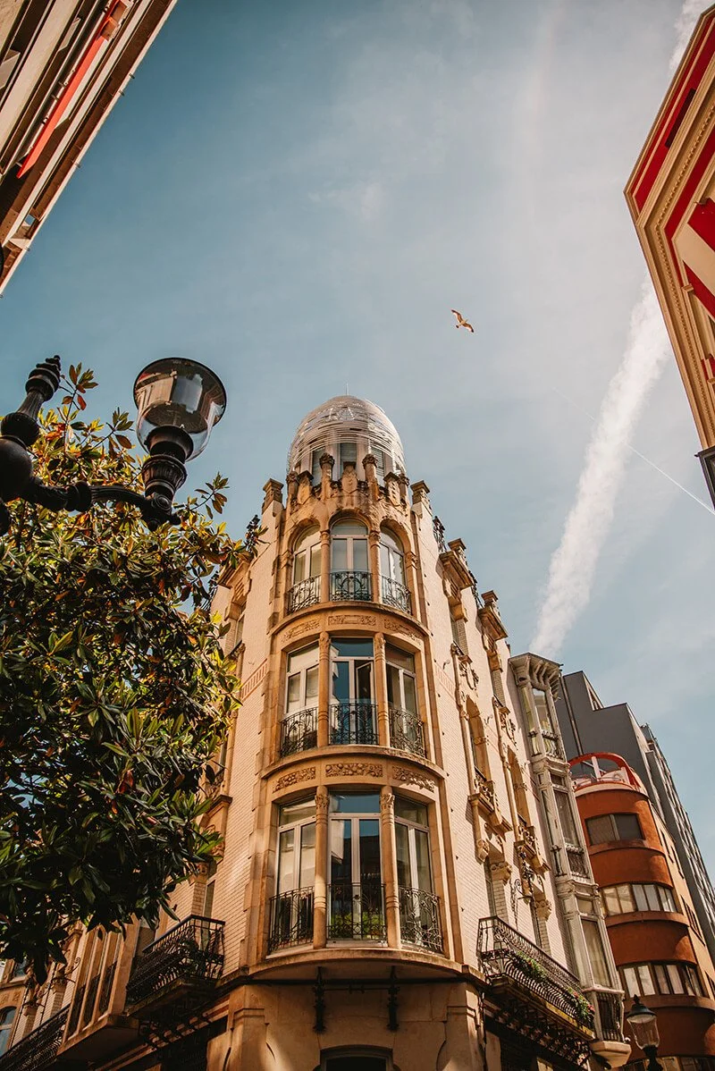 A tall, ornate building with rounded balconies and decorative columns, viewed from below against a blue sky with a bird flying and a contrail. Shot in Gijon, Asturias, Spain.