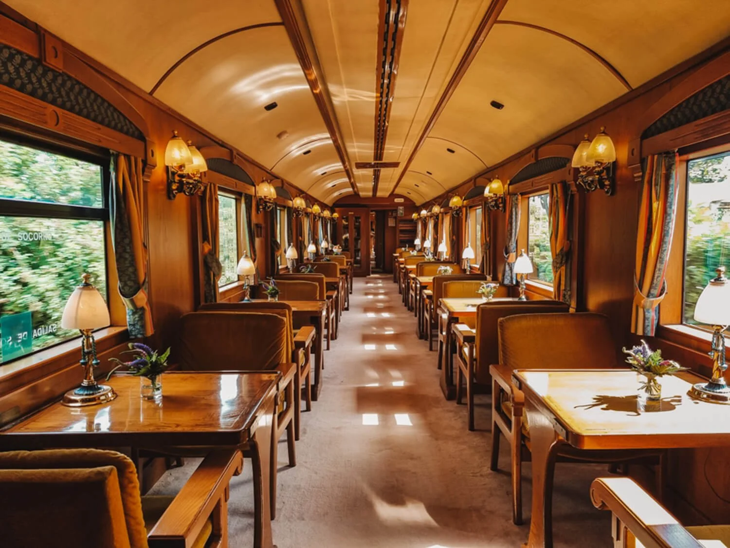 Interior of a vintage train dining car of the Costa Verde Express in Northern Spain, with wooden tables, upholstered chairs, table lamps, and windows with curtains, decorated in warm wood tones.