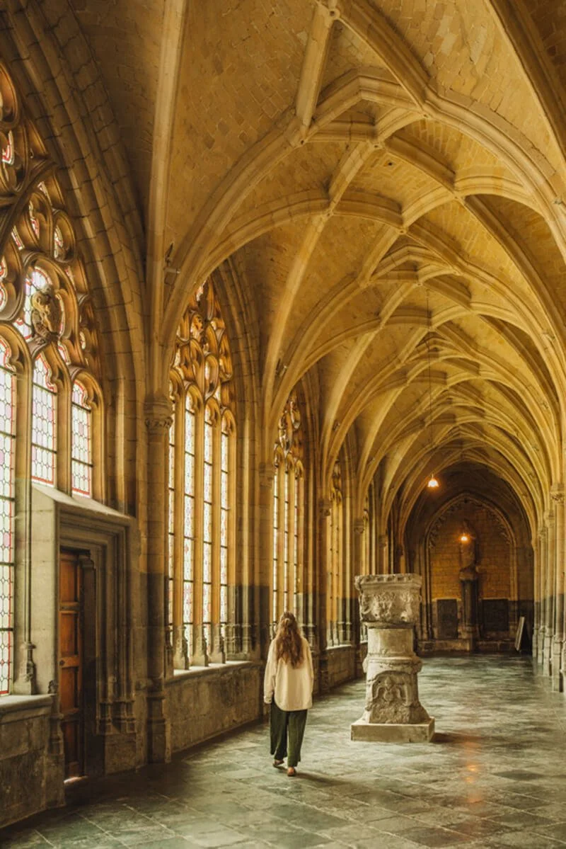 Woman walking through a large, ornate cathedral corridor with stained glass windows and vaulted ceilings. Shot in Liège, one of the most beautiful places in Belgium.