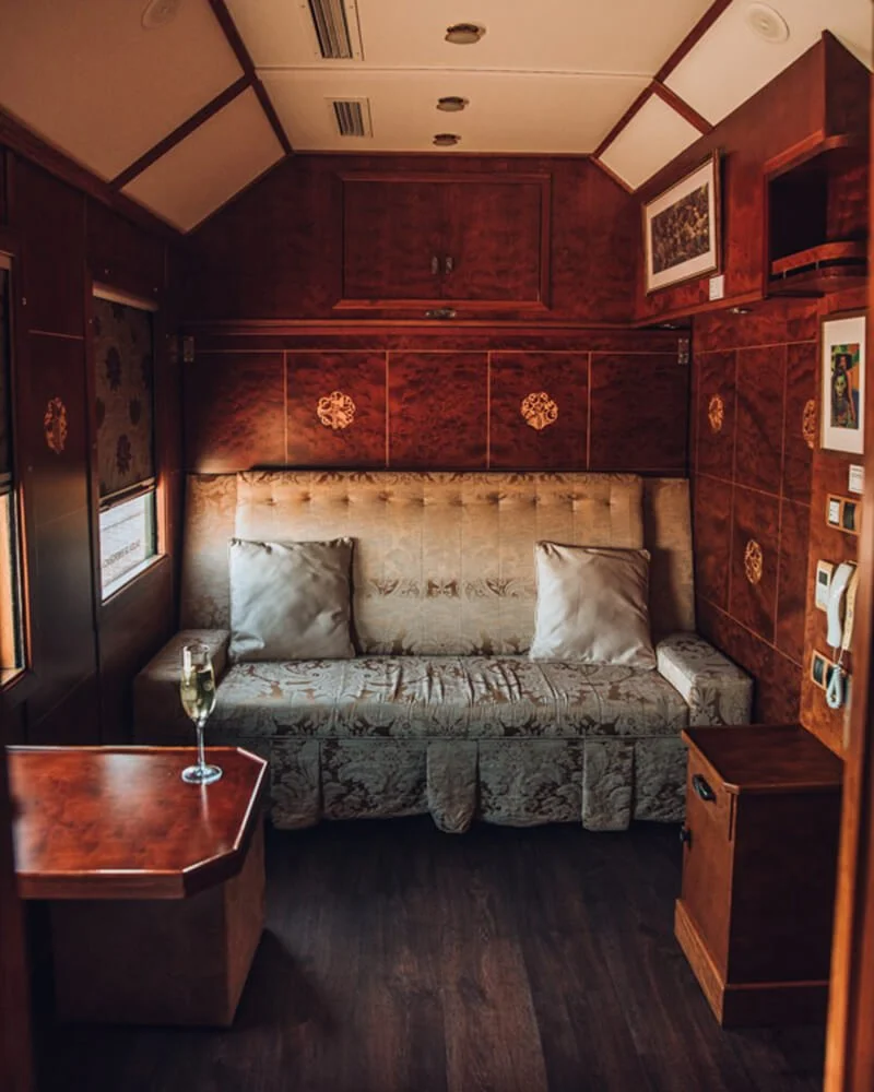 Small living room with wooden walls, a floral patterned sofa, two cushions, a small wooden side table, and a window with floral curtains. This is the cabin of the Al Andalus train in Southern Spain.