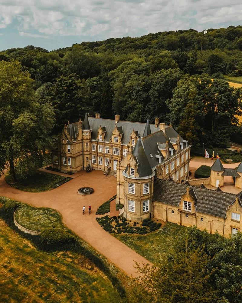 A large historic castle with multiple towers and turrets, surrounded by lush green trees and a winding pathway leading up to it, in a scenic countryside setting. One of the many castles in Belgium.