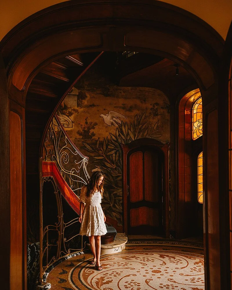 A young woman in a white dress standing on a decorative circular carpet in an ornate hallway with wooden walls, a curved staircase, and stained glass windows. Art Nouveau in Brussels, Maison Hannon.