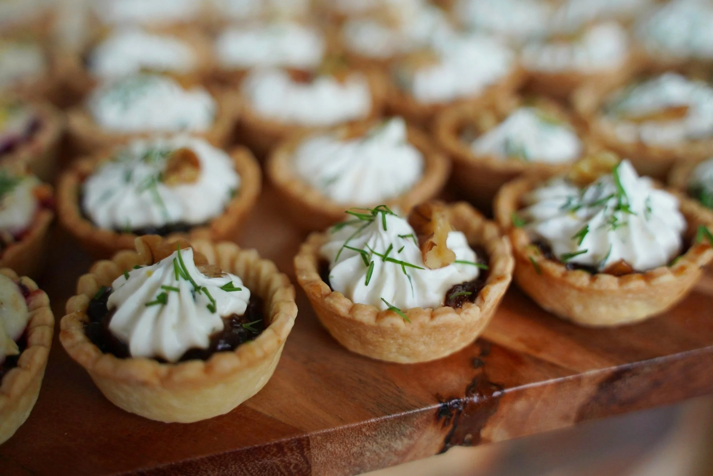 Mini savory tartlets with whipped cream, garnished with herbs, on a wooden serving board.