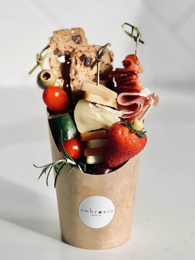 A paper cup filled with various appetizers including cherry tomatoes, strawberries, cheese, sliced meats, olives, and bread with chocolate chips.