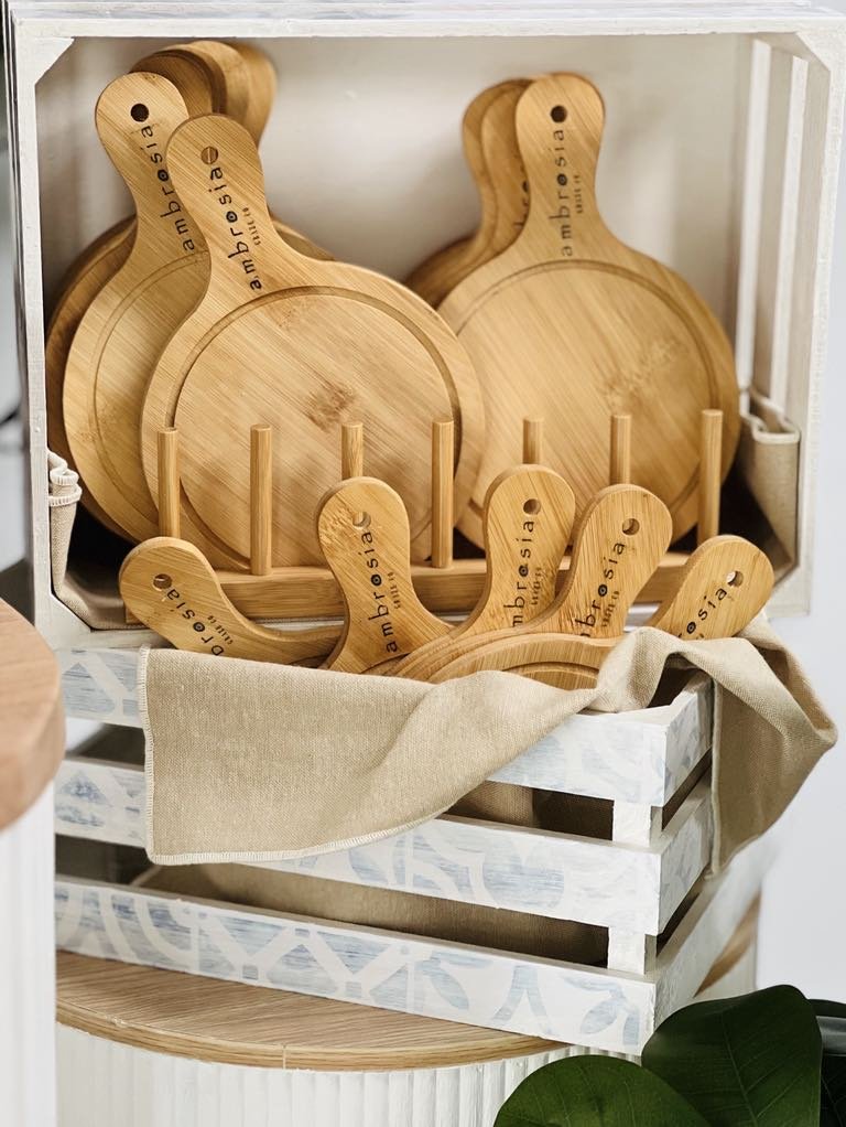 Set of round wooden cutting boards with handles, stored on a white wooden shelf and a basket, with the Instagram handle '@ambrosia' engraved on the handles.