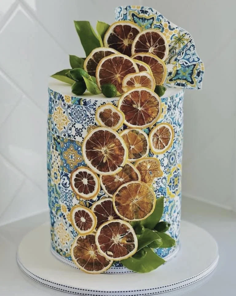 Decorative cake with dried citrus slices and green leaves, wrapped in colorful patterned paper and placed on a white cake stand.