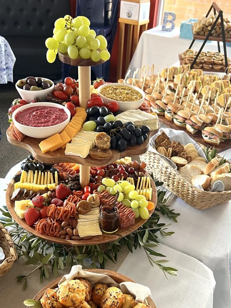 A three-tiered cheese and fruit platter with grapes, strawberries, cheeses, olives, cherry tomatoes, crackers, and various spreads at a party or gathering table.