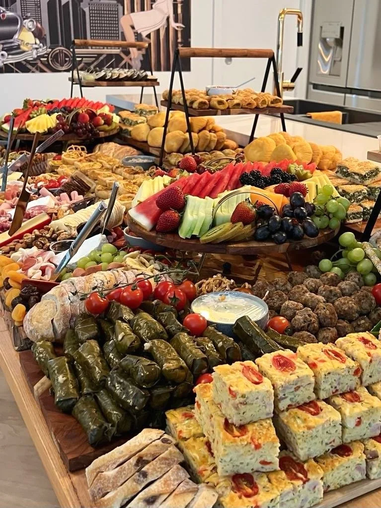 A buffet table with an assortment of fresh fruit, pastries, stuffed grape leaves, cherry tomatoes, and various finger foods and desserts.