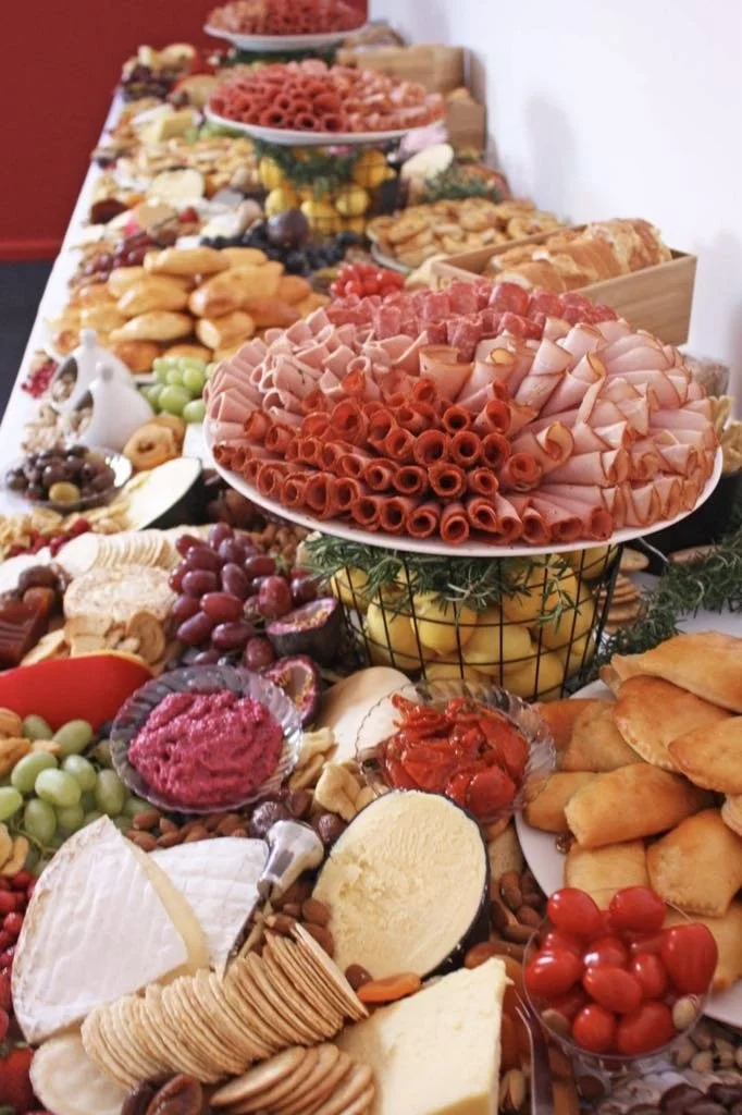 A table filled with an assortment of foods, including sliced meats, cheeses, fruits, crackers, grapes, tomato salad, cookies, and various other snacks for a buffet or party.