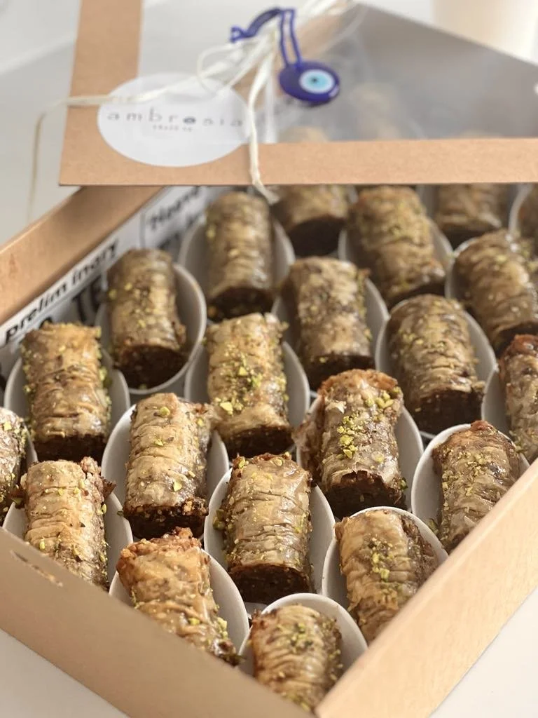 A box filled with small, rolled pastries topped with crushed pistachios, each placed in individual white paper cups.