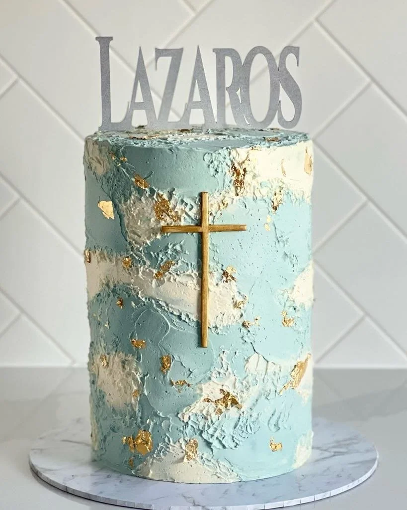 A tall, two-tone blue and white textured cake with gold accents, a gold cross on the front, and a topper that reads 'Lazaros' in silver letters.