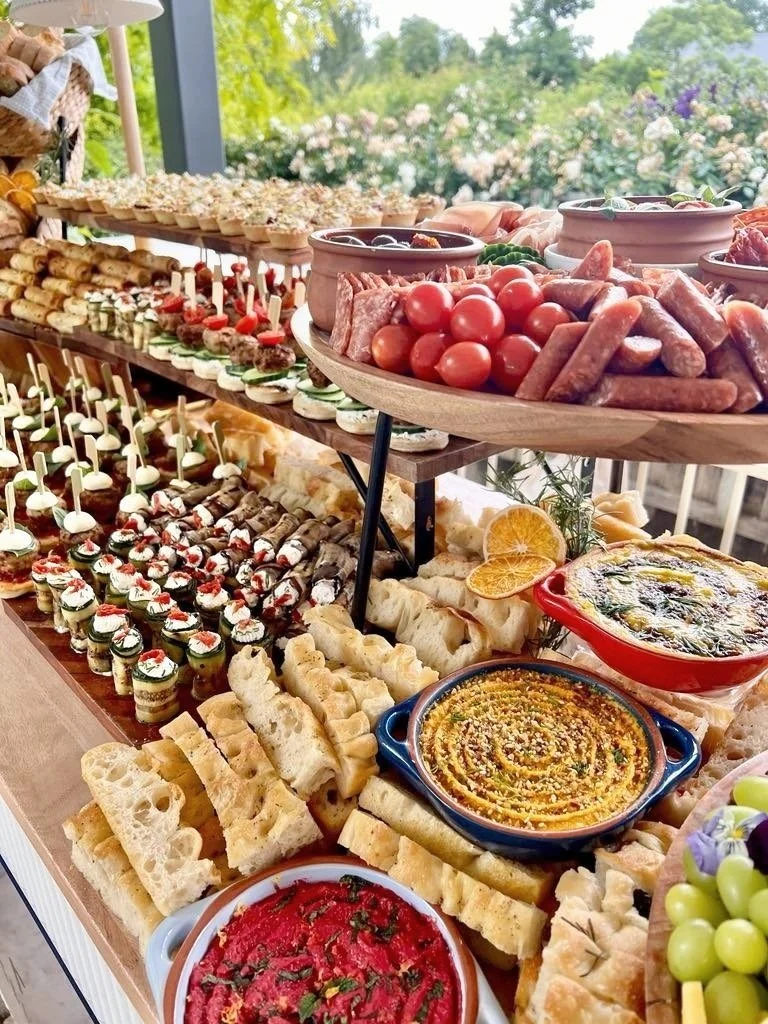 An outdoor buffet table filled with assorted appetizers, dips, finger foods, cherry tomatoes, sausages, mini sandwiches, skewered snacks, and fresh vegetables, set against a scenic garden background.