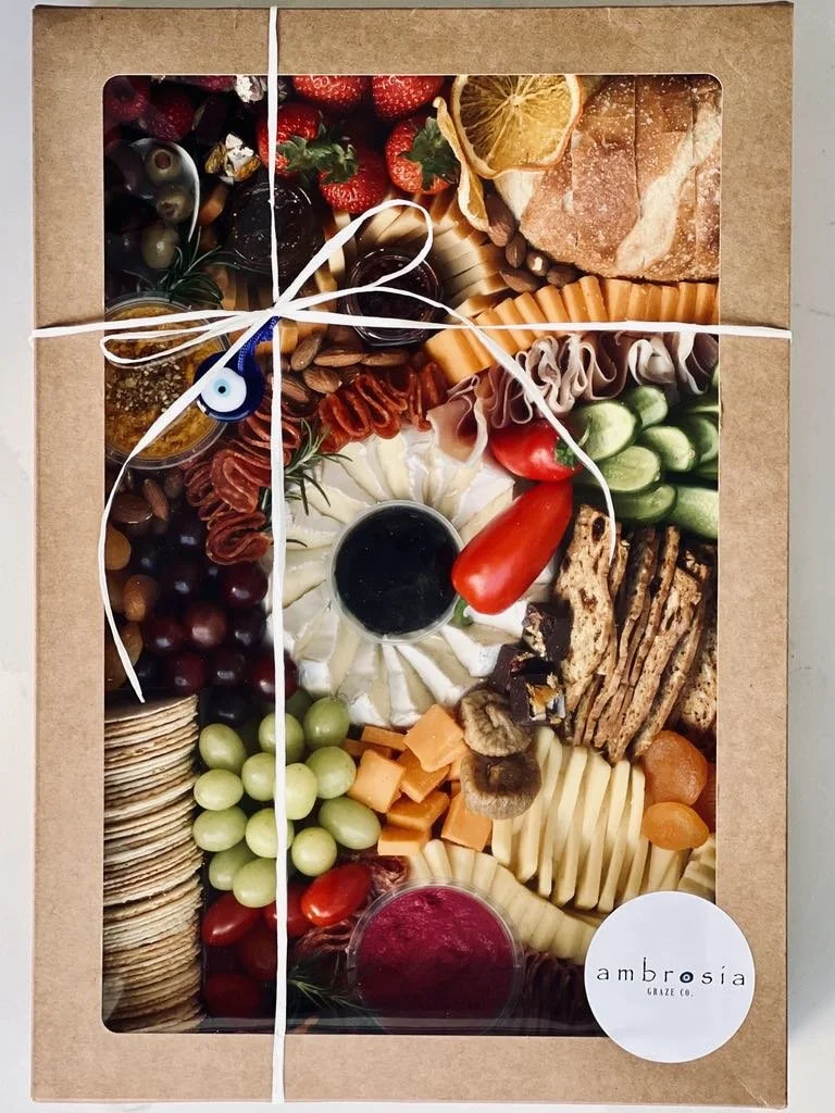 Box of assorted cheeses, crackers, grapes, strawberries, cherry tomatoes, sliced vegetables, cured meats, dried fruits, honey, and a container of jam, all arranged for a charcuterie platter, wrapped with white string and labeled 'ambrosia'.