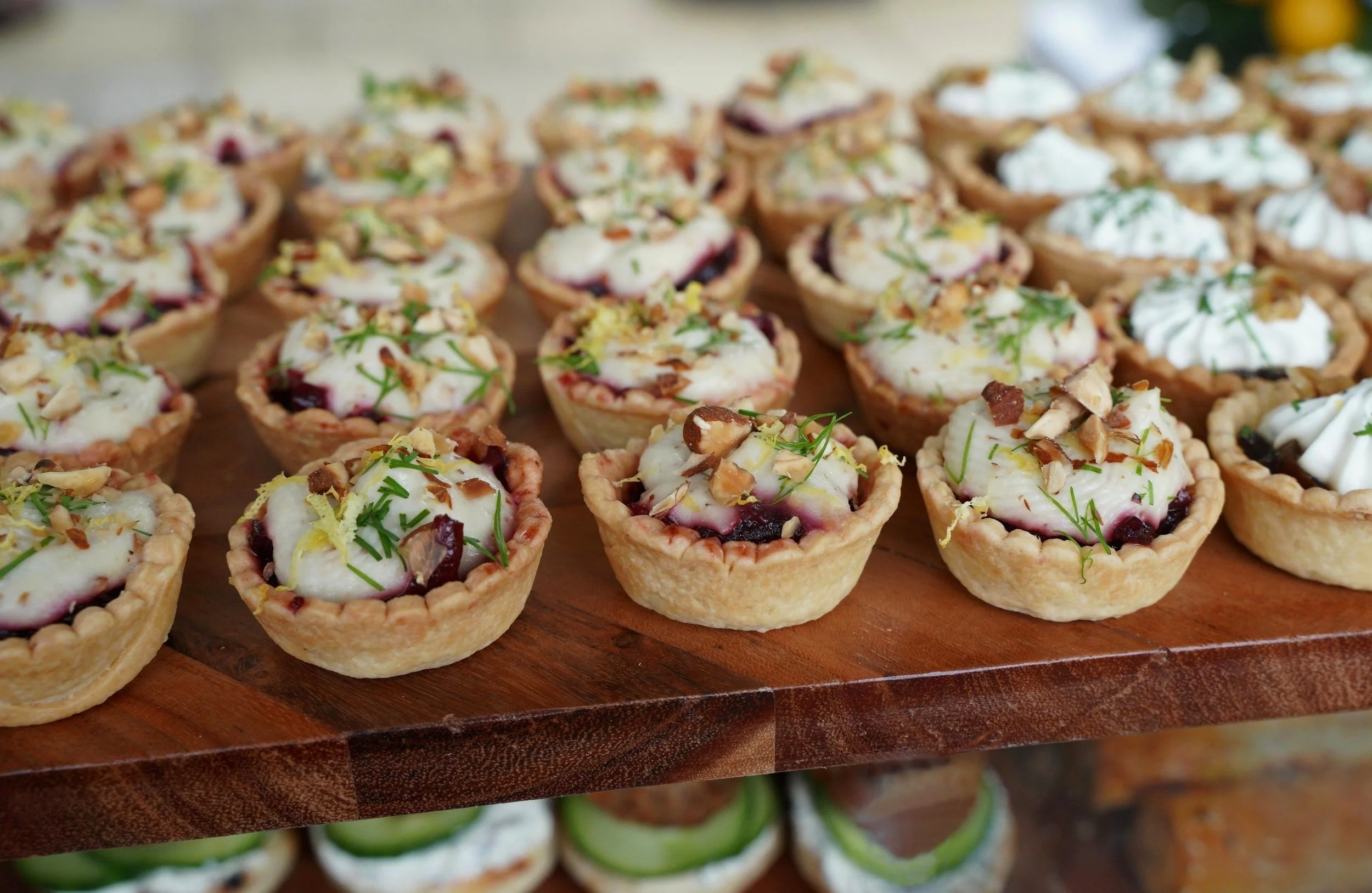 Assorted mini savory tarts with toppings like herbs, cheese, and chopped nuts on a wooden serving tray.